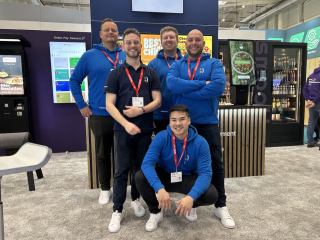 Fünf Männer posieren am Messestand, vier stehen und einer kniet vorne; mehrere tragen blaue Hoodies mit Logo und rote Messeausweise, im Hintergrund sind Bestell- und Automaten-Displays zu sehen.