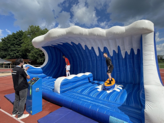 Outdoor-Event mit aufblasbarer Surf-Wellenbahn: zwei Personen stehen in der blauen Wellenkulisse, eine Person bedient daneben das Steuerpult.