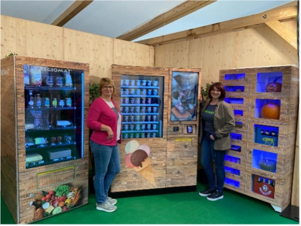 Zwei Frauen stehen in einem Holzraum vor mehreren REGIOMAT-Verkaufsautomaten mit beleuchteten Produktfächern und einem zentralen Automaten mit Bildschirm und Zahlmodul.