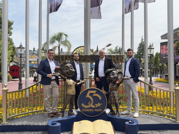 Vier Männer stehen vor dem Eingang des Europa-Park neben zwei Maskottchen-Figuren und einem ‚50 Jahre Europa-Park‘-Schild, im Hintergrund Blumenbeete und Parkkulisse.