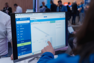 Nahaufnahme eines Monitors mit der Actindo-Core1-Oberfläche ‚Workflows‘; eine Person zeigt mit dem Finger auf ein Prozessdiagramm, im Hintergrund sind Messebesucher unscharf zu sehen.