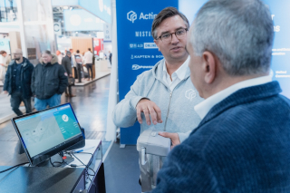 Zwei Männer im Gespräch an einem Messestand; einer demonstriert ein Gerät/Terminal neben einem Bildschirm, im Hintergrund sind Besucher und ein Actindo-Banner zu sehen.