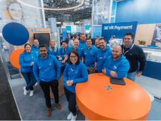 Gruppenfoto am VR-Payment-Messestand: mehrere Mitarbeitende in blauen Hoodies stehen und lächeln vor dem VR-Payment-Logo, mit orangefarbenem Stehtisch im Vordergrund.