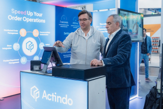 Zwei Männer stehen an einem Actindo-Messestand und bedienen ein POS-Terminal; im Hintergrund ist eine Präsentationswand mit ‚Speed Up Your Order Operations‘ zu sehen.