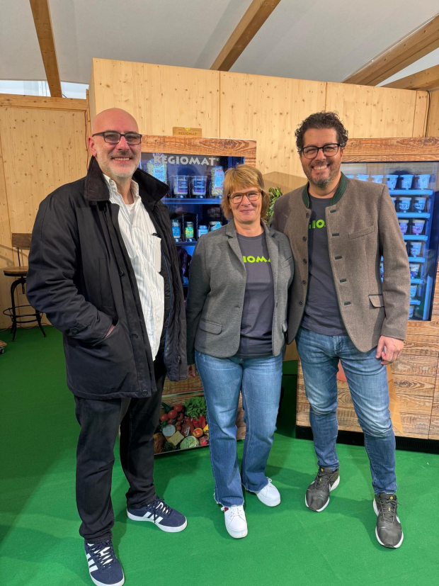 Drei Personen stehen lächelnd in einem Holzraum vor einem REGIOMAT-Automaten mit regionalen Produkten; im Hintergrund sind Regale mit Waren sichtbar.
