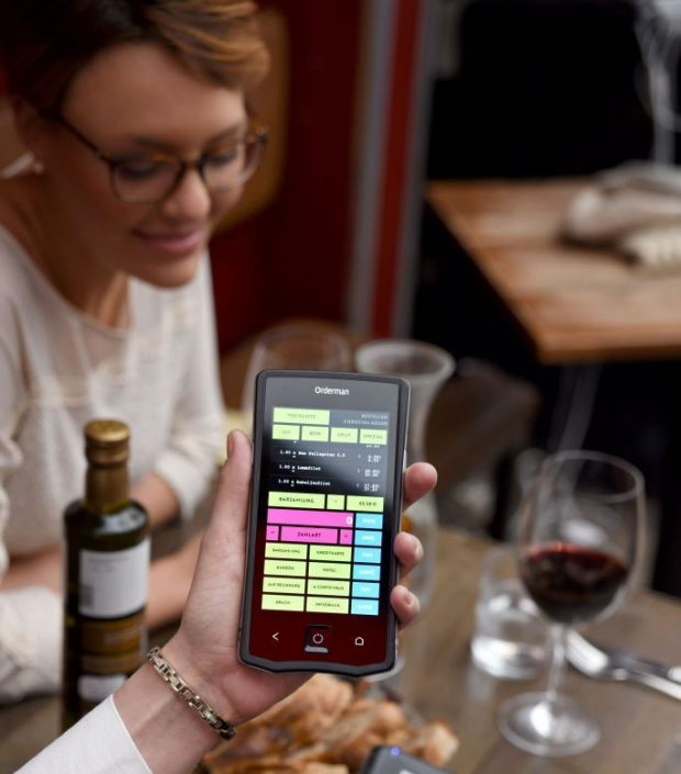 Hand hält ein mobiles Orderman-Kassengerät mit Bestell- und Zahlungsübersicht; im Hintergrund sitzt eine Frau am Tisch mit Wein und Gläsern.